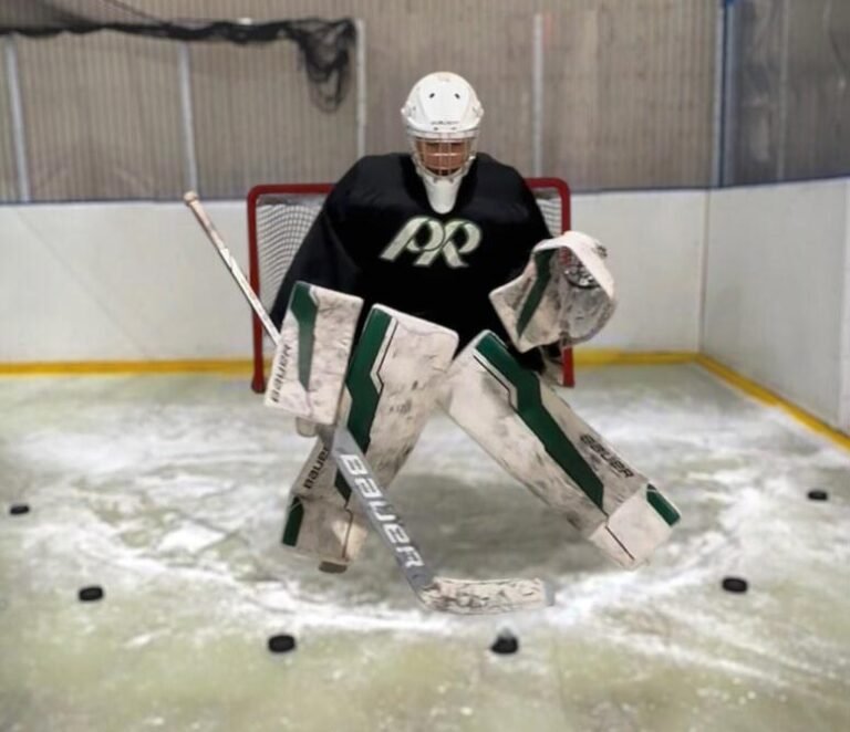 Youth hockey goalie in full gear mid-save in front of a red net at an indoor training rink, Bauer pads and stick, pucks on the synthetic ice around the crease